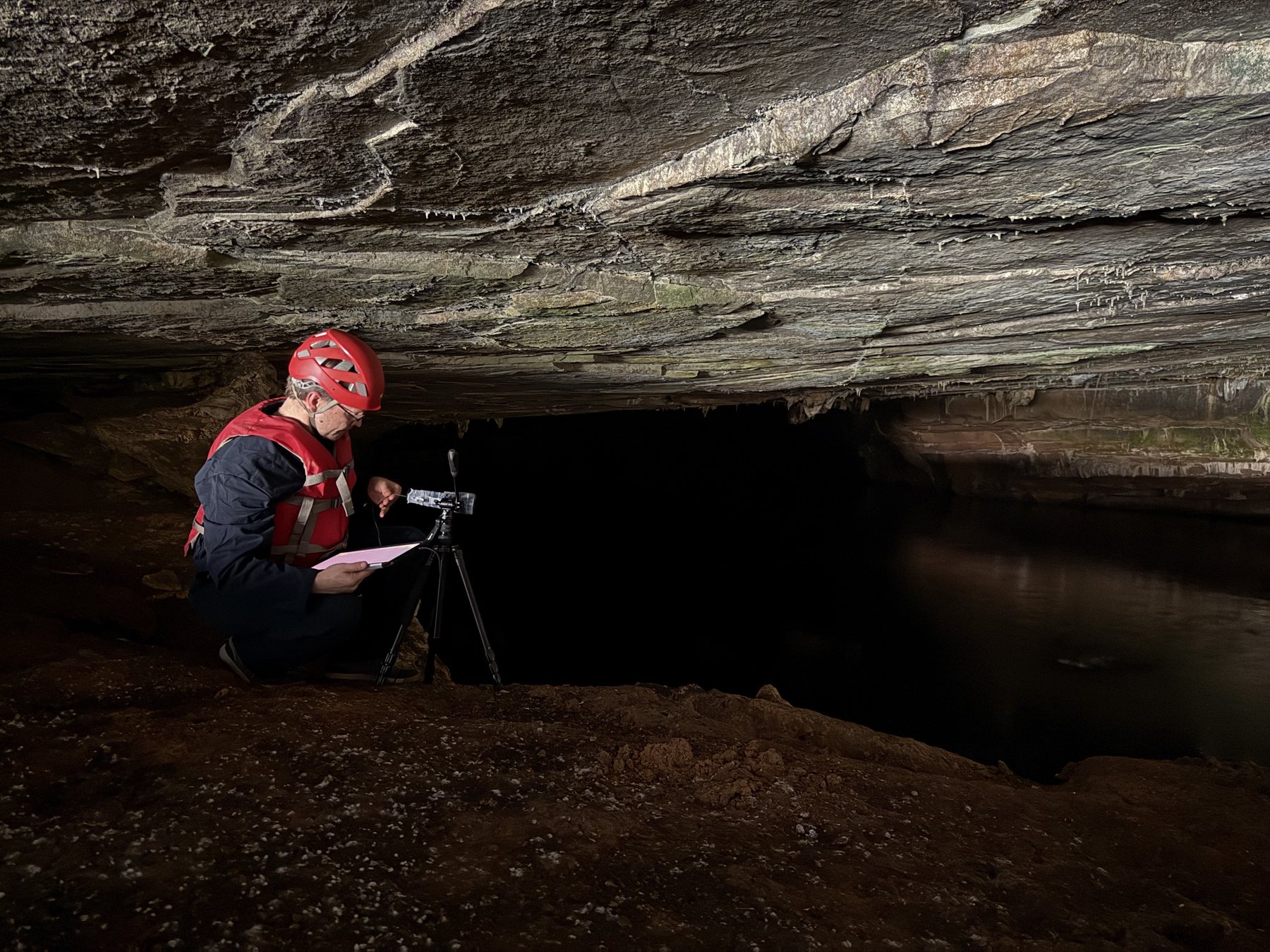 A researcher in a helmet setting up a camera on a tripod at a cave entrance
